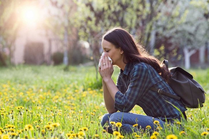 Young pretty woman blowing nose in grassland with spring flowers. Pollen allergy symptoms