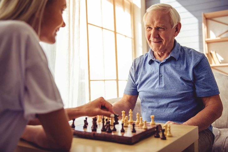 Beautiful young girl-volunteer and handsome old man are playing chess and smiling