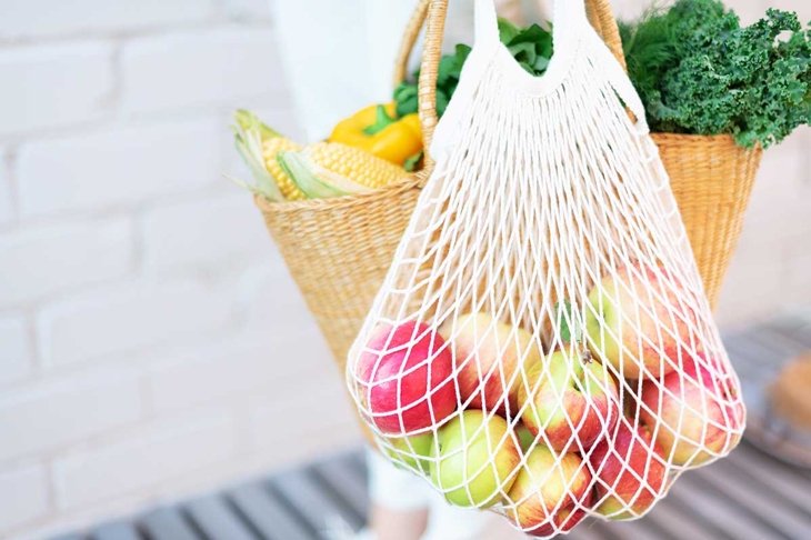 Zero waste concept with copy space. Woman holding straw basket and reusable mesh shopping bag withapples, vegetables, white brick background. Eco friendly mesh shopper. Copy space.