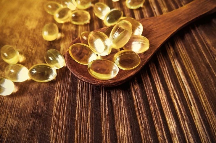 Vitamin D yellow pills on wood spoon on wooden background & soft sun light. Medicine gel tablets of vitamin d on table. Yellow vitamin D oil capsule keeps healthy while lack of sun - medicine concept