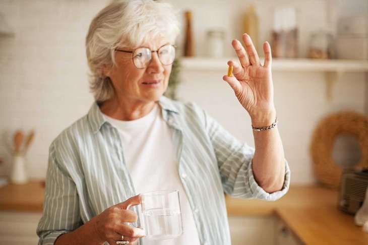 Beautiful mature sixty year old female in stylish eyeglasses holding mug and omega 3 supplement capsule, going to take vitamin after meal. Senior gray haired woman taking fish oil pill with water