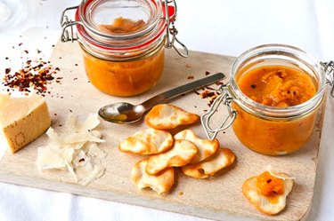 Jars of English peach and ginger chutney on wooden board with Parmesan and chips.
